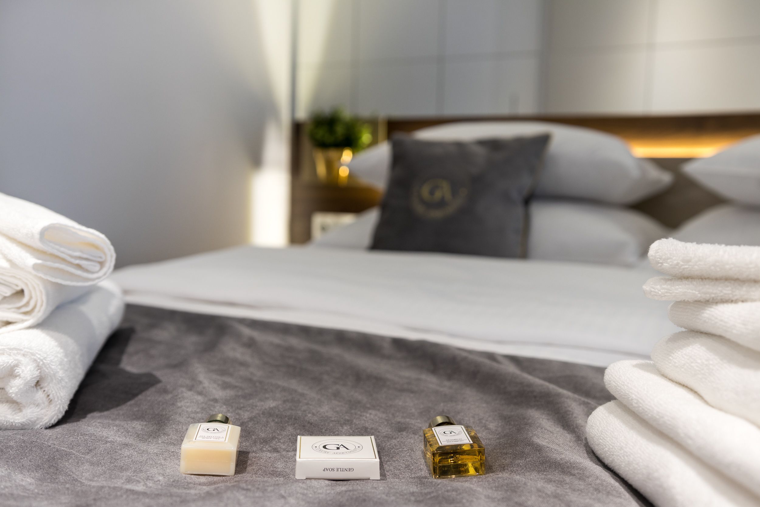 Un lit bien fait, avec du linge de lit blanc, une couverture grise et deux articles de toilette portant la lettre « G » sur une surface grise.