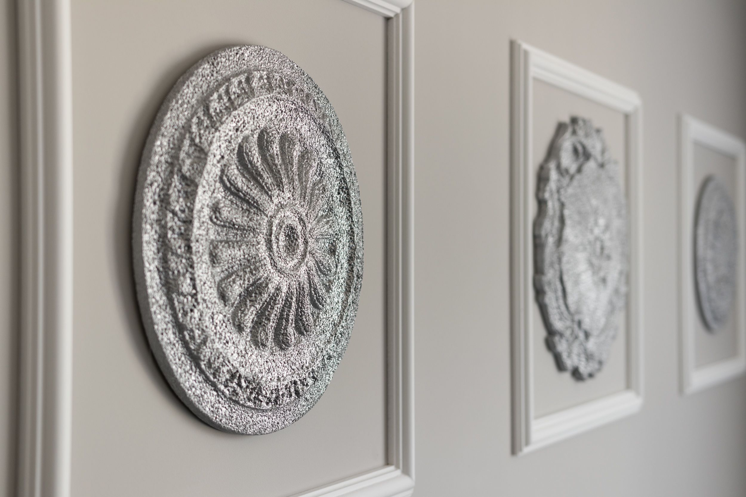 Un mur orné de tableaux encadrés, dont une œuvre circulaire à motifs floraux et une œuvre rectangulaire à la surface texturée.