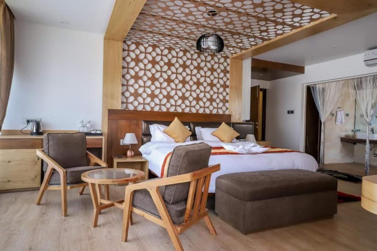 A modern hotel room with a large bed, two wooden chairs, a small round table, and a wooden ceiling with a geometric pattern.