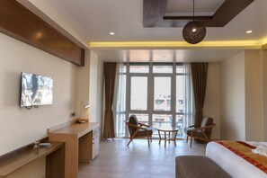 Et moderne hotelværelse med en seng, skrivebord, to stole, et lille bord og et stort vindue med gardiner.