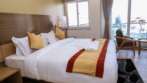 A neatly made bed with white bedding and a red and yellow accent, a wooden headboard, a bedside table with a lamp, and a view of the city through large windows. - Retro Hotel and Spa (Kathmandu)
