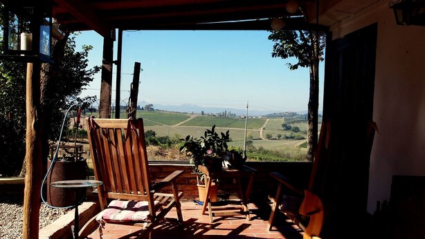 A patio with a wooden chair, a table, and a potted plant. A view of a green landscape with fields and mountains is visible through the open space. - Agriturismo La Terra (Montepulciano)