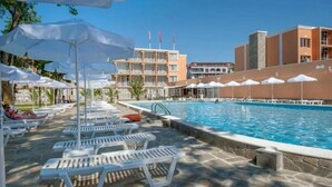 Zona de piscina con sillas de descanso y sombrillas, rodeada de edificios con balcones.