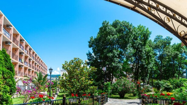 Un sendero conduce a un edificio con balcones, rodeado de abundante vegetación y un cielo azul despejado.
