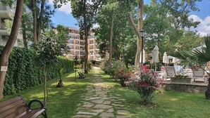 Un sendero de piedra que conduce a un banco y zona de descanso rodeados de vegetación y un edificio al fondo.