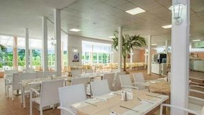 Un comedor interior amplio con mesas y sillas blancas, ventanas grandes y una planta en el centro.