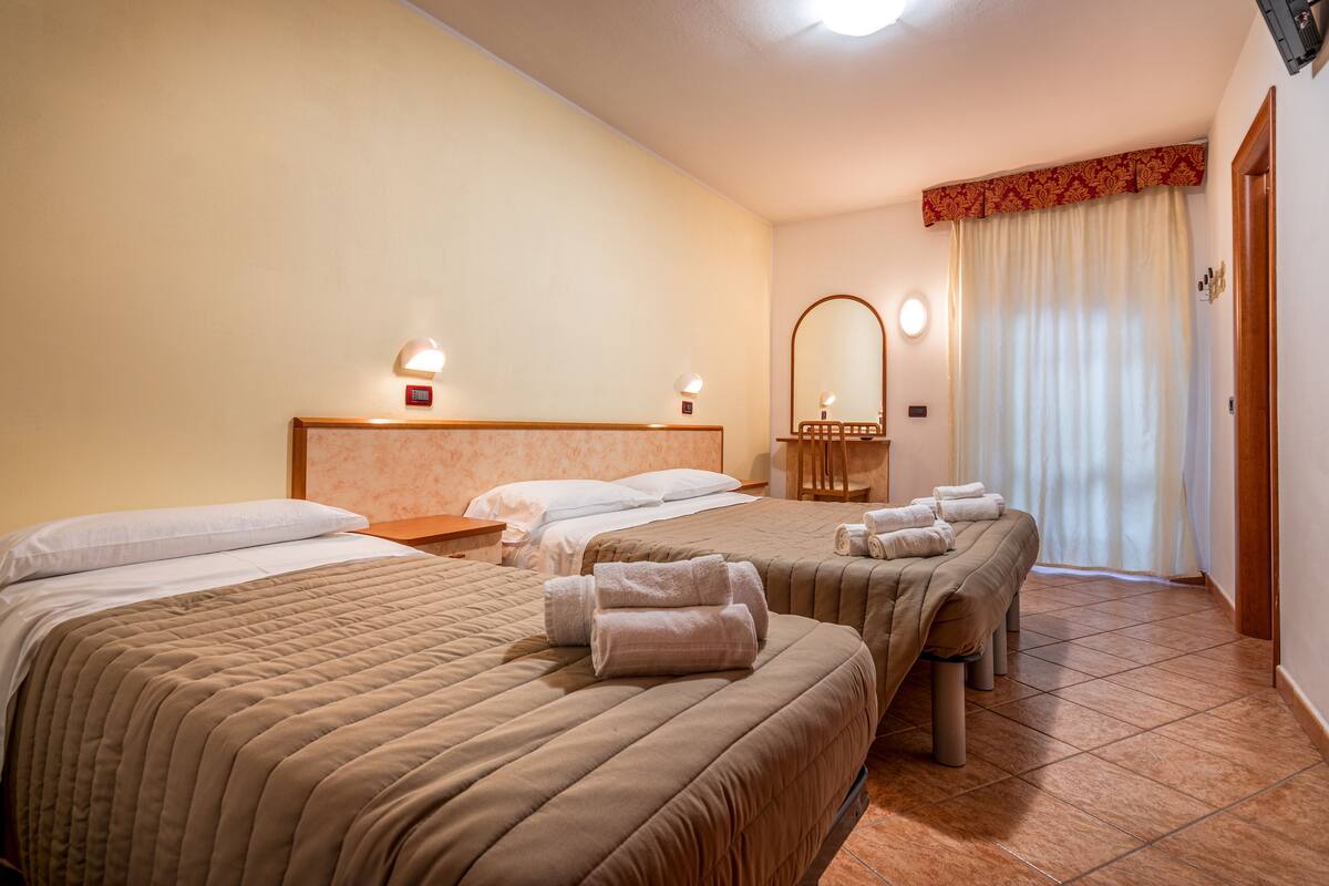 A hotel room with three beds, each with a brown quilted bedspread and white pillows. There are two bedside tables with lamps, a large mirror above the headboard, and a window with a curtain.