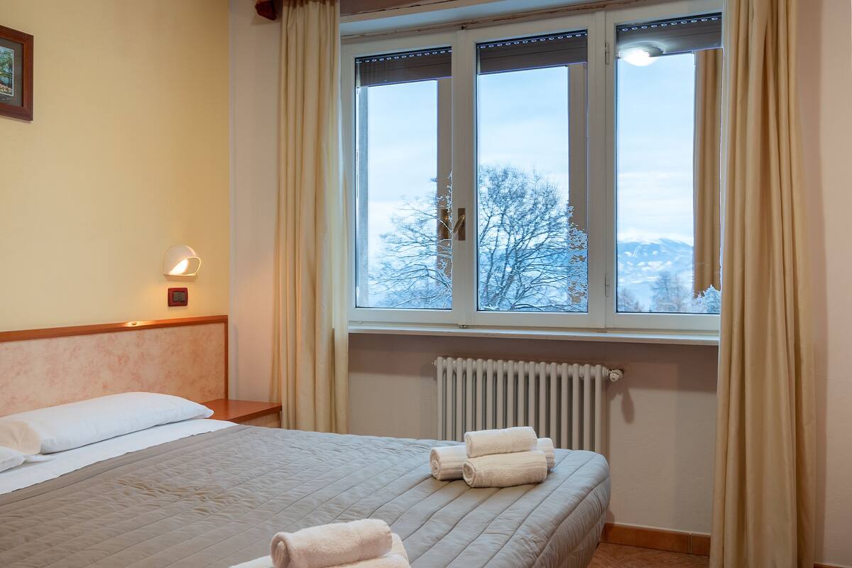 A hotel room with a bed, a window with curtains, a radiator, and a wall-mounted lamp.