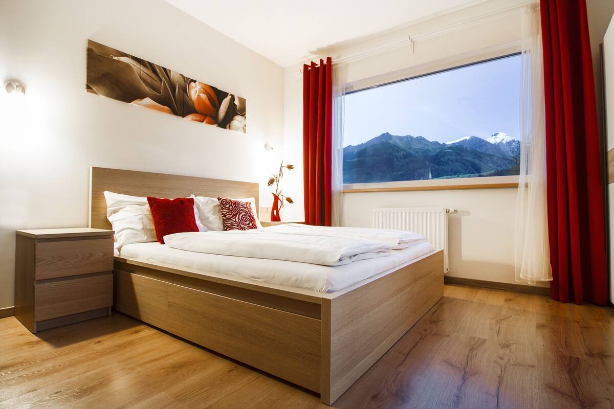A modern bedroom with a wooden bed, a nightstand, a window with a mountain view, and a painting on the wall.