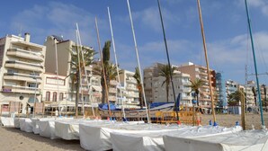 Una playa con barcos cubiertos por sábanas blancas, palmeras y edificios de varios pisos al fondo.