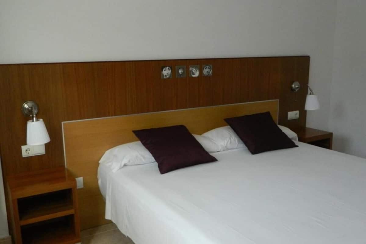 A hotel room with a wooden headboard, two pillows, a white bedspread, and two bedside tables with lamps.