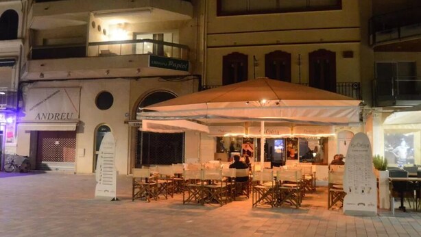 Un café al aire libre con mesas y sillas bajo un paraguas grande, rodeado de edificios con tiendas y un letrero que dice "ANDREU".