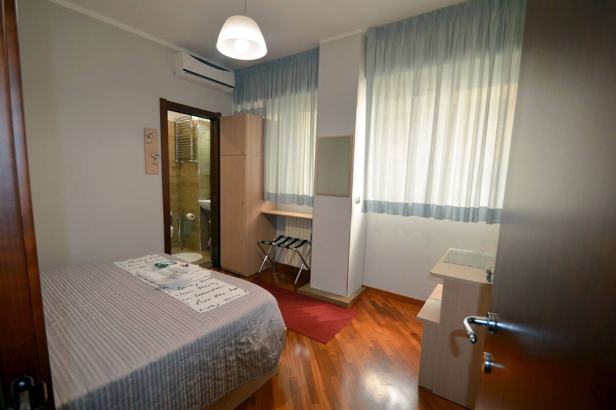 A hotel room with a bed, a desk, a mirror, and a window with curtains.