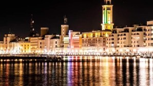 A nighttime cityscape with illuminated buildings and a water fountain reflecting lights.
