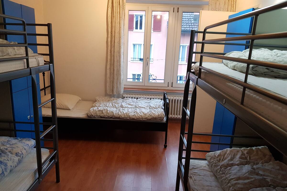 A dormitory room with bunk beds, a wooden floor, and a window with a view of buildings outside.