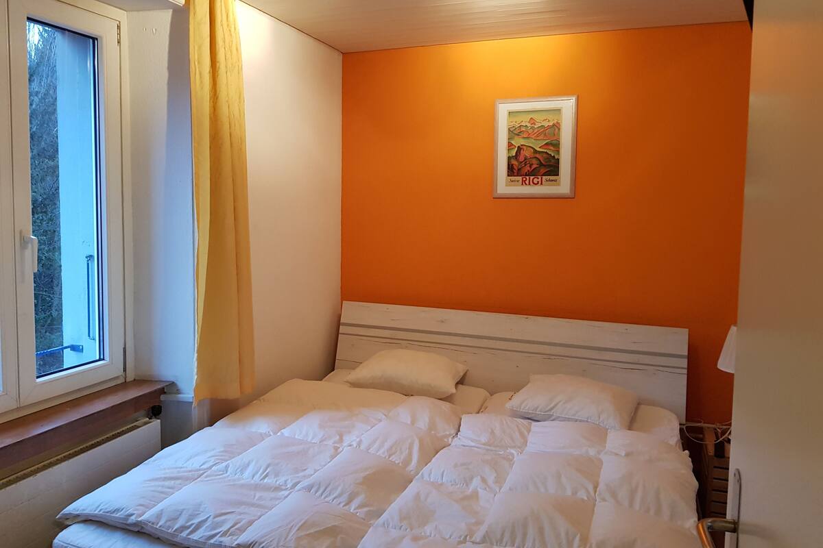 A bedroom with a bed, a window with curtains, an orange wall, and a framed picture.