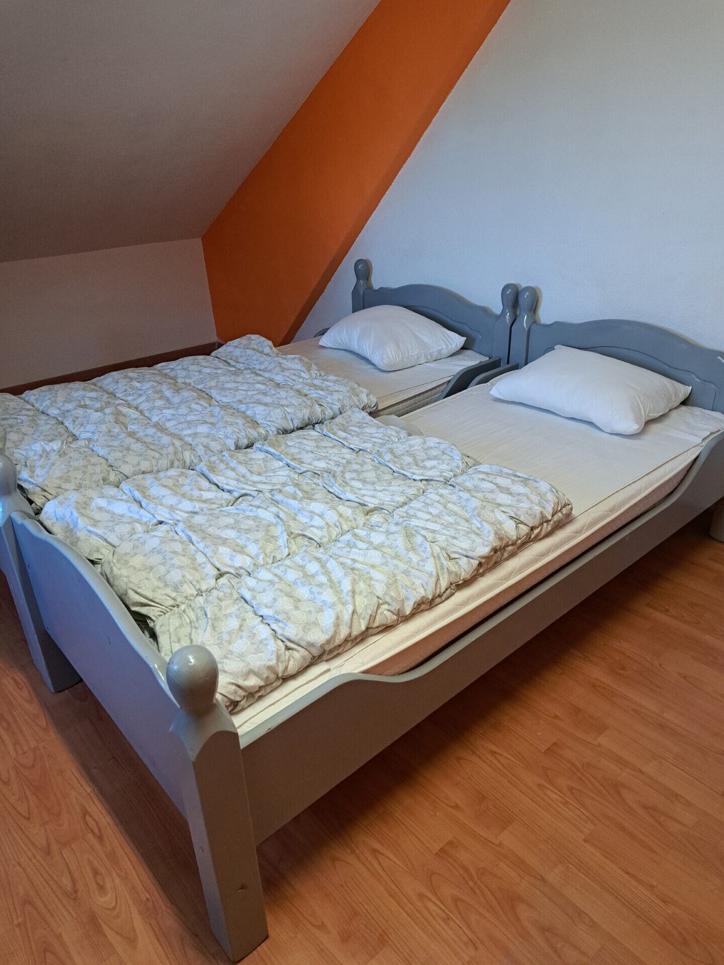 Un lit simple avec un cadre en bois et une couette à motifs, dans une pièce au plancher en bois et dont un mur est peint dans une teinte orange.