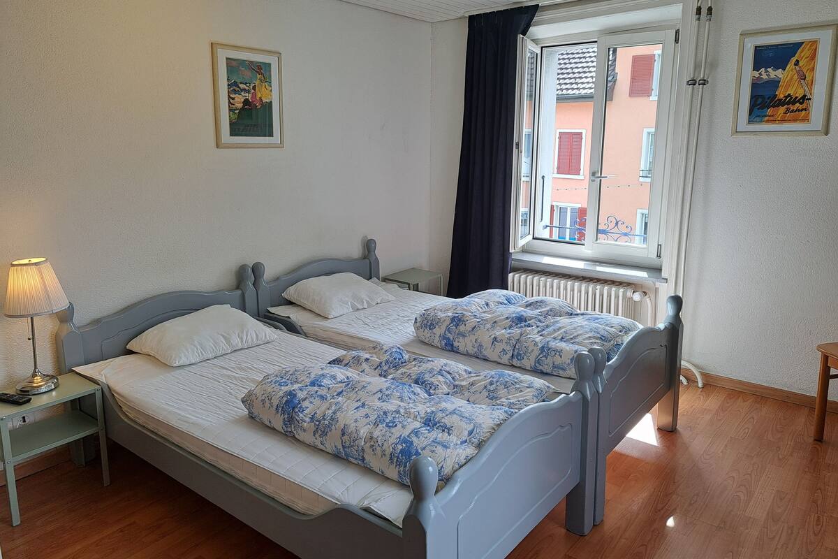A double bed with floral bedding, a bedside lamp, and a window with a view of buildings.
