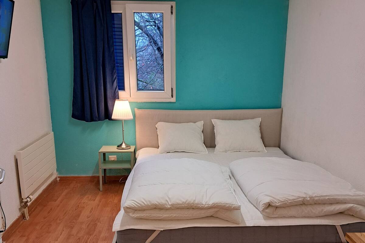 A bedroom with a bed, a nightstand, a window with curtains, and a radiator.