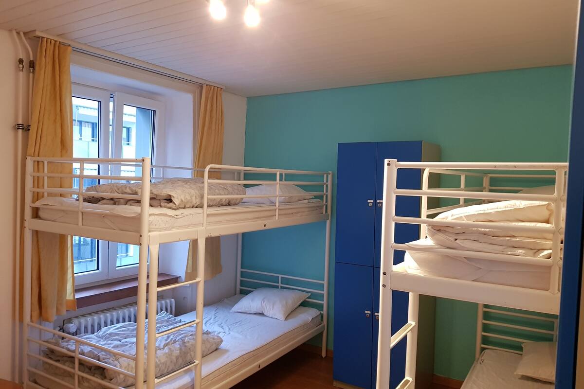 A dormitory room with bunk beds, a radiator, a blue wardrobe, and a window with curtains.
