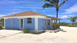 Un pequeño edificio de una sola planta con fachada azul y techo marrón, ubicado en una playa arenosa con una palmera cerca.