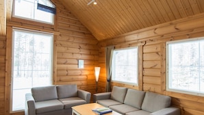 A log cabin interior with two grey sofas, a wooden table, and large windows offering a view of a snowy forest.