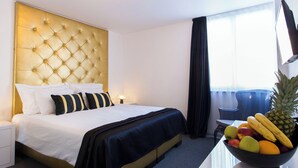 Una habitación de hotel con cabecera capitonada, cama con ropa de cama blanca y negra, frutero en la mesita de noche y ventana con cortinas.