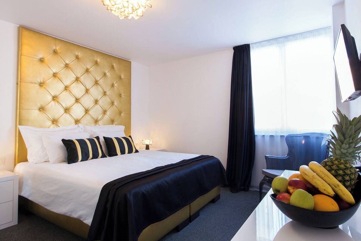A hotel room with a tufted headboard, a bed with white and black bedding, a fruit bowl on the nightstand, and a window with curtains.