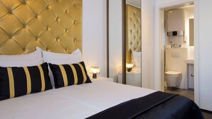 Una habitación de hotel con cabecera capitonada, un espejo grande, una cama con ropa de cama blanca y negra, y un baño visible con inodoro y lavabo.