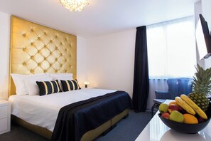 Ein Hotelzimmer mit einer gepolsterten Kopfstütze, einem Bett mit weißer und schwarzer Bettwäsche, einer Obstschale auf dem Nachttisch und einem Fenster mit Vorhängen.