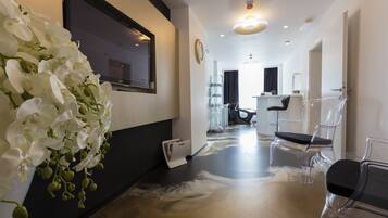 A modern interior with a hallway leading to a salon area, featuring a large mirror, clear chairs, and a decorative orchid arrangement.