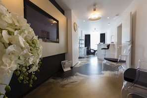 A modern interior with a hallway leading to a salon area, featuring a large mirror, clear chairs, and a decorative orchid arrangement. - B Gold Bandb (Split)