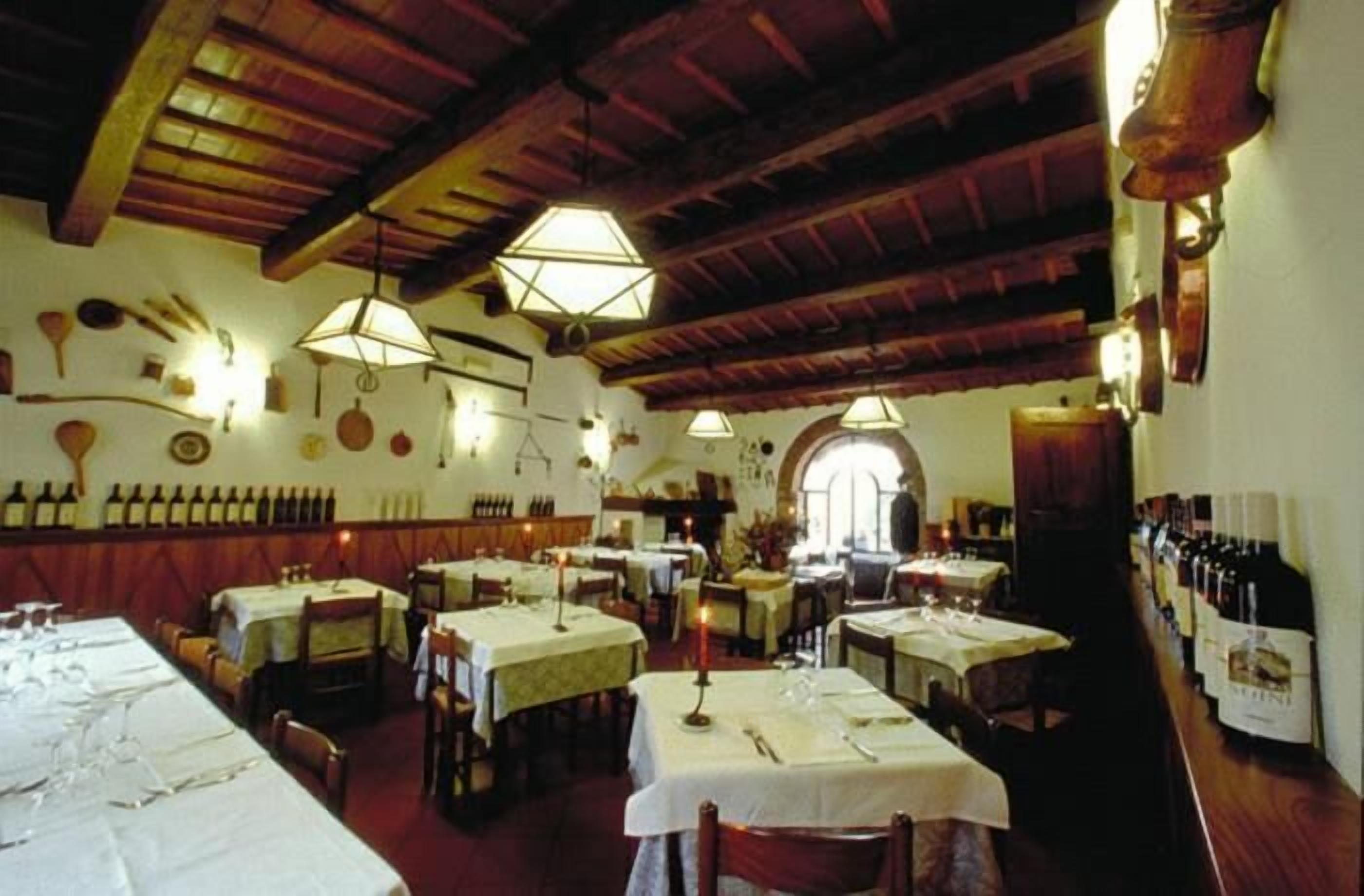 A restaurant with wooden beams, hexagonal chandeliers, and wine bottles on the wall. Tables are set with white tablecloths and candles.