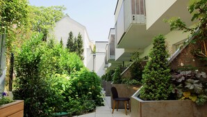 Um pátio urbano estreito com muita vegetação, um banco de madeira e um edifício moderno com varandas.