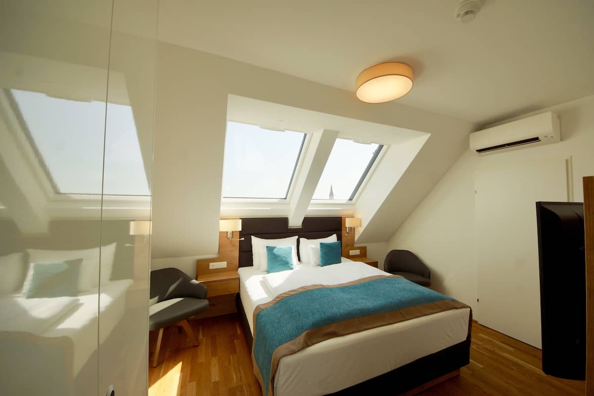 Um quarto moderno com claraboia, uma cama com colcha azul e castanha, uma mesinha de cabeceira, uma cadeira e um aparelho de ar condicionado.