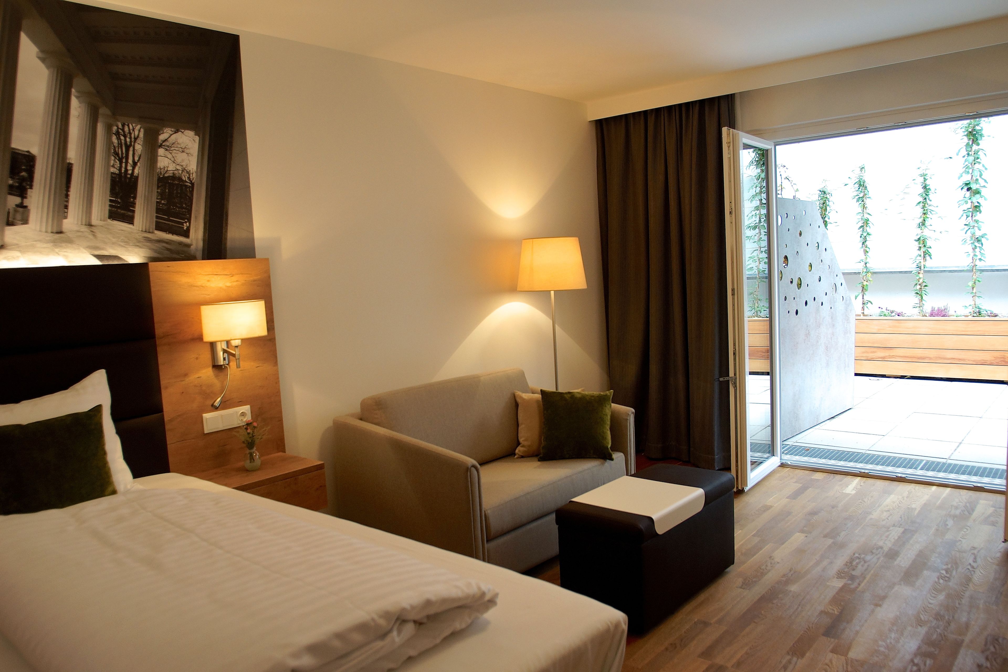 A hotel room with a bed, a sofa, a small table, and a lamp. There is a window with curtains and a view of greenery outside.