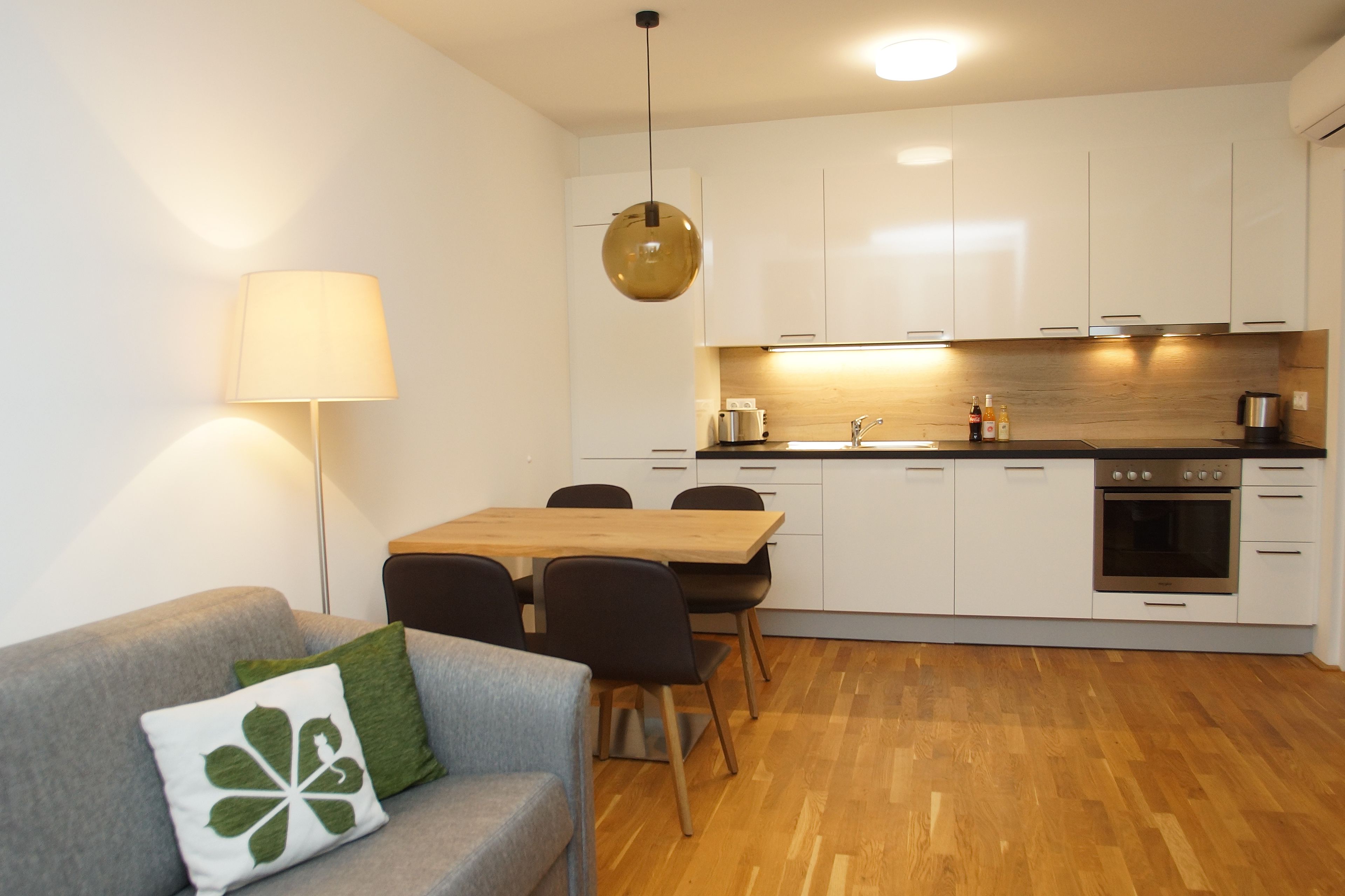 Una cocina moderna con armarios blancos, encimera de madera y horno empotrado. Hay una mesa de comedor con sillas y una lámpara.