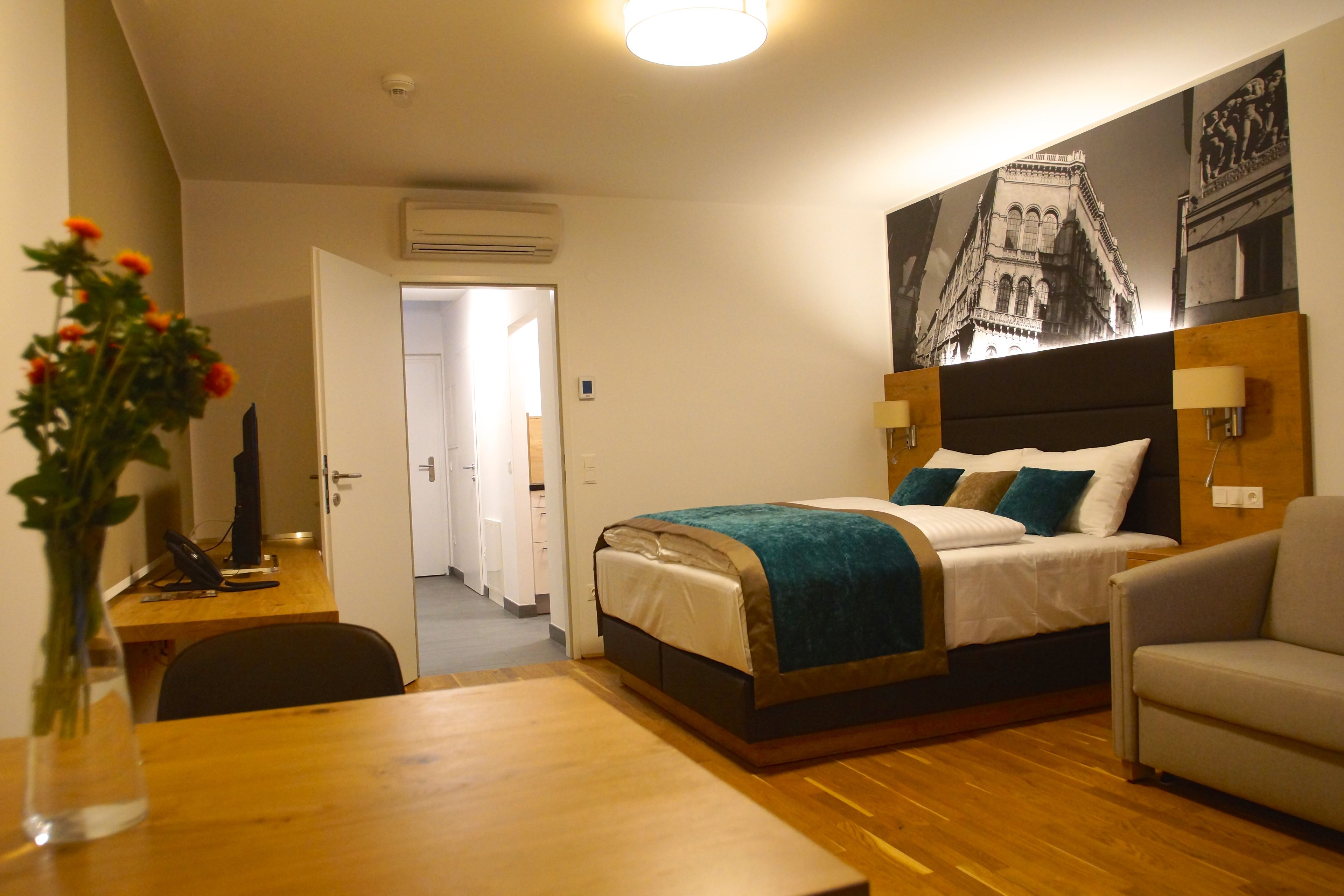 A hotel room with a bed, a desk, a chair, a sofa, a vase with flowers, and a wall-mounted lamp.
