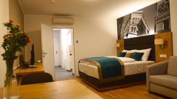 A hotel room with a bed, a desk, a chair, a sofa, a vase with flowers, and a wall-mounted lamp.
