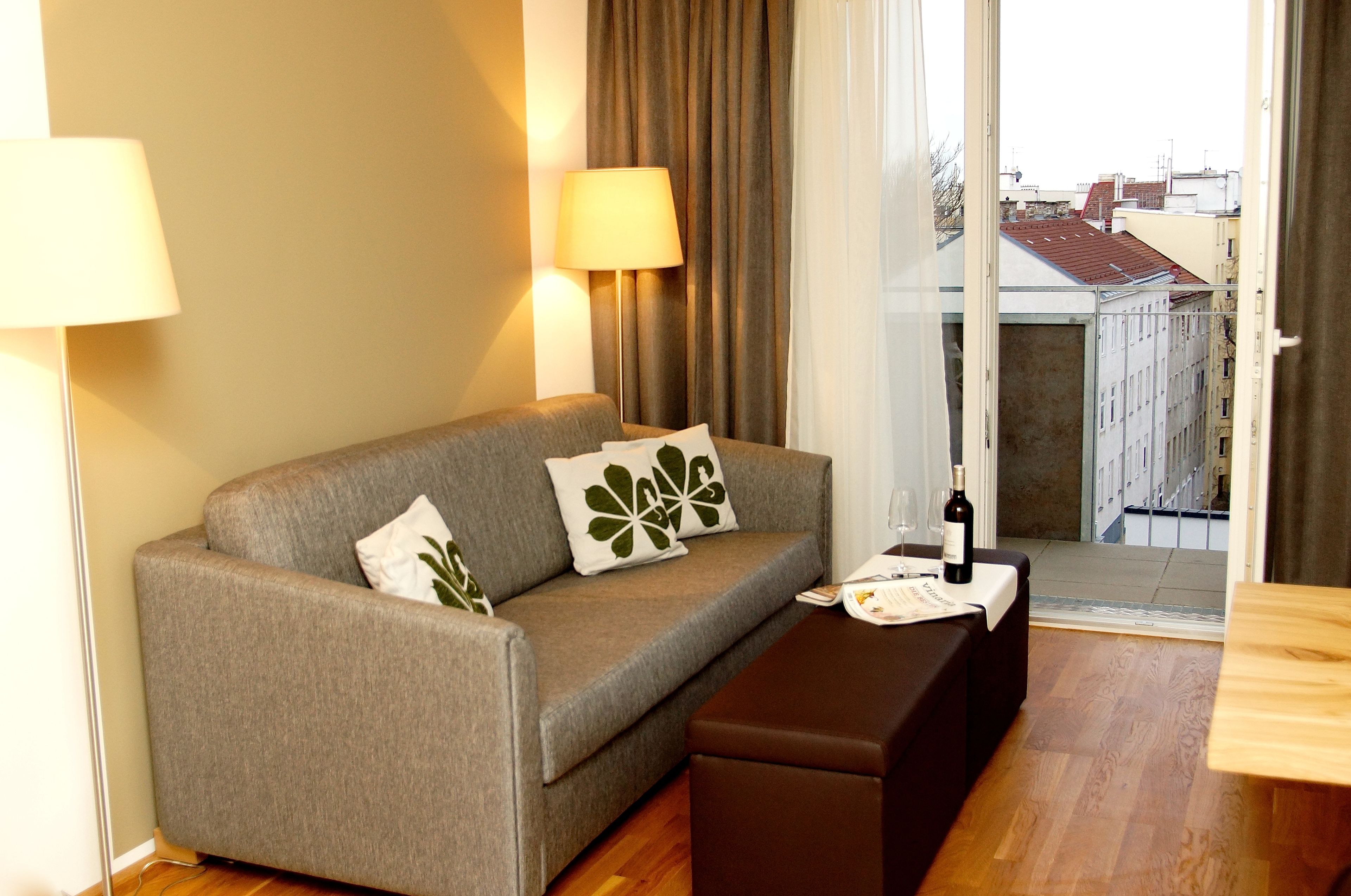 Une chambre d’hôtel moderne avec un canapé gris, un banc en bois, une lampe et une vue sur la ville, visible à travers une grande fenêtre.