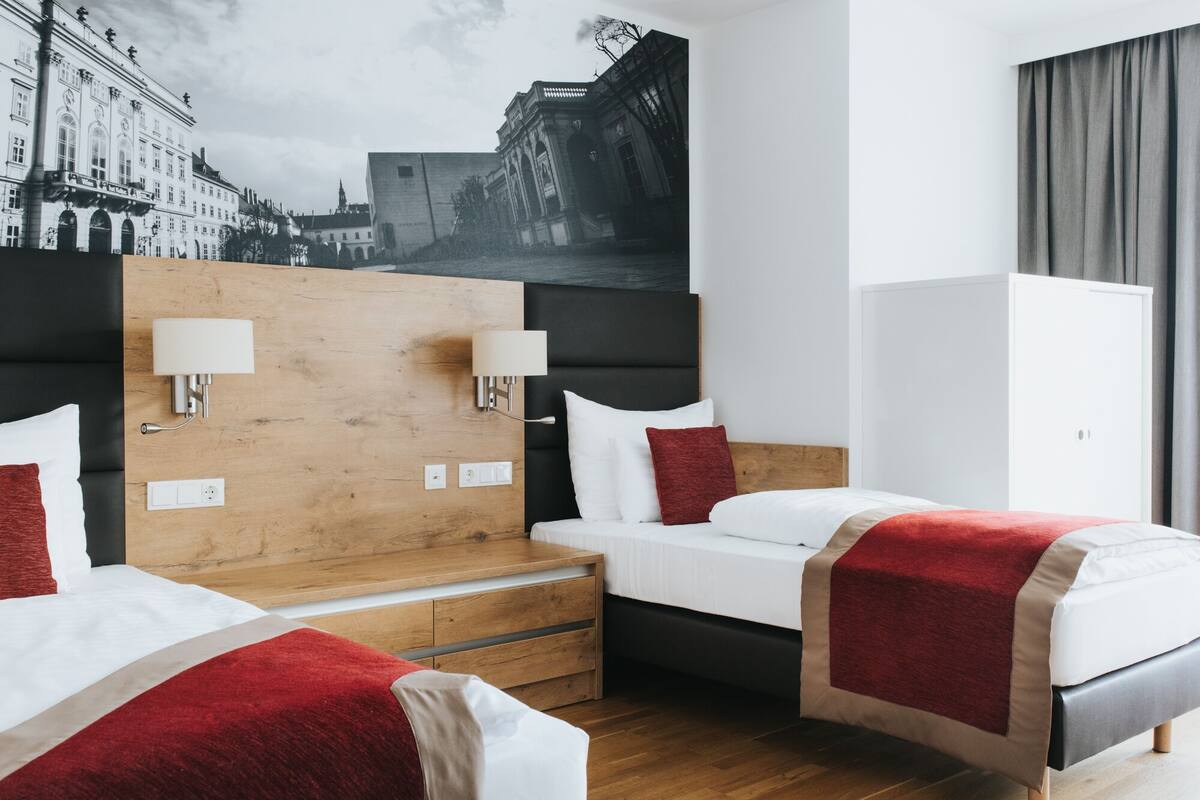 Quarto de hotel com duas camas, cabeceira de madeira, parede branca e uma grande fotografia em preto e branco de uma paisagem urbana.
