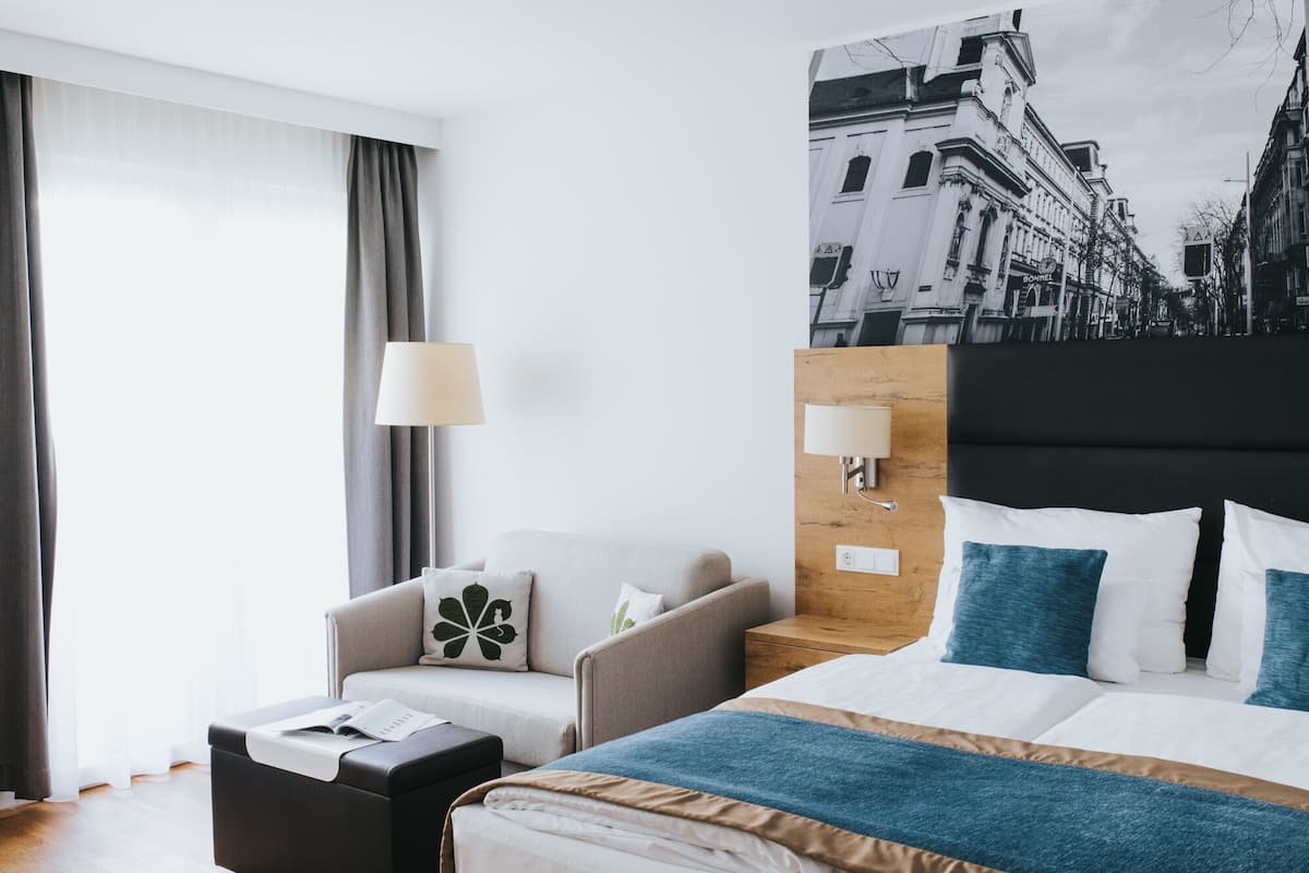 Quarto de hotel com cama, duas poltronas, mesa de apoio, luminária e uma grande pintura de paisagem urbana na parede.