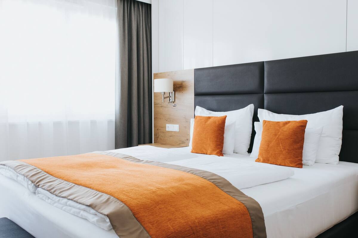Quarto de hotel com uma cama grande, cabeceira, mesa de cabeceira, luminária e janela com cortinas.