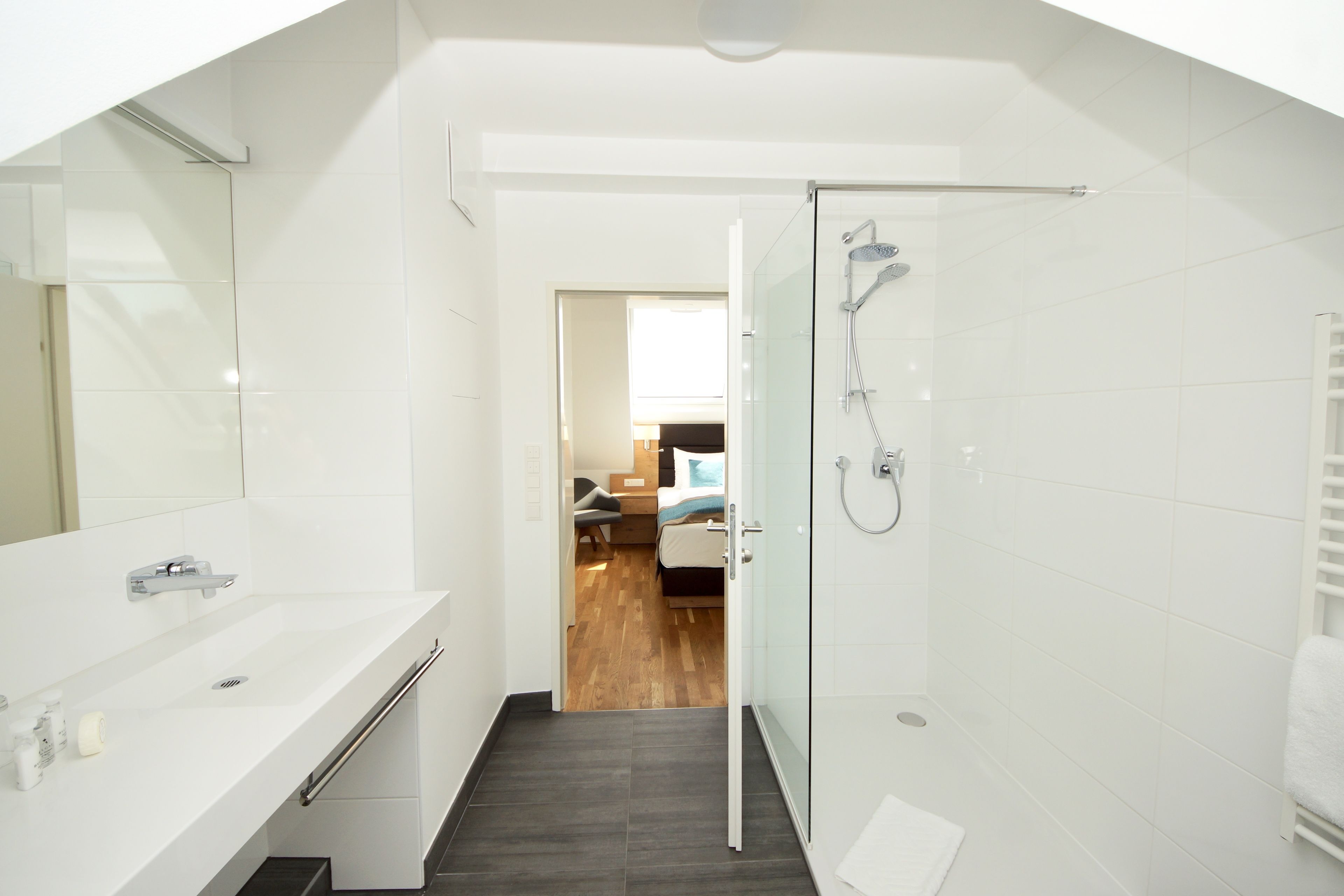 Un baño moderno con cabina de ducha de vidrio, un lavamanos blanco y una habitación visible al fondo.