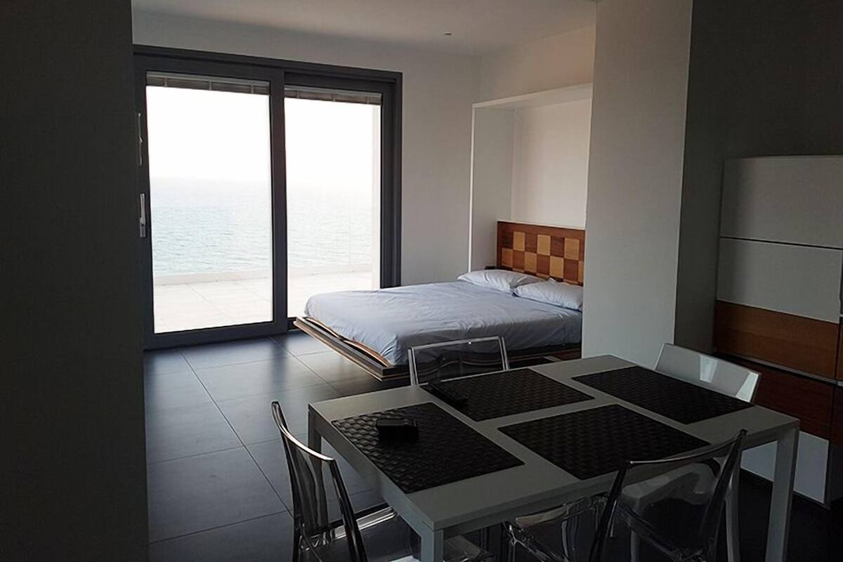 Une chambre d’hôtel moderne, équipée d’un lit, d’une table à manger avec des chaises et offrant une vue sur la mer grâce à de grandes fenêtres.