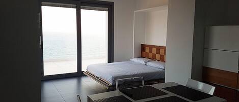 A modern hotel room with a bed, a dining table with chairs, and a view of the sea through large windows.