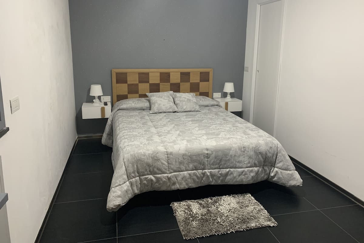 Une chambre à coucher bien rangée, avec une tête de lit à carreaux, deux tables de chevet avec des lampes, une couvre-lit grise à motifs et un petit tapis sur le sol.