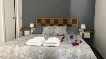 Letto rifatto con cura, con copripiumino grigio fantasia, due cuscini e asciugamani piegati. Sul comodino, un boccale di vetro con un fiore.