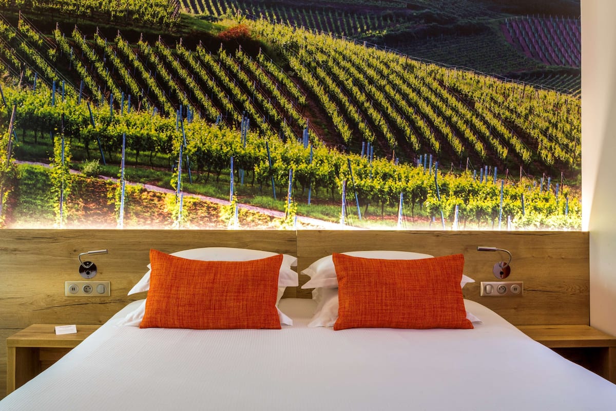 Habitación de hotel con una cama grande, dos almohadas naranjas, un cabecero de madera y vistas a un viñedo por la ventana.