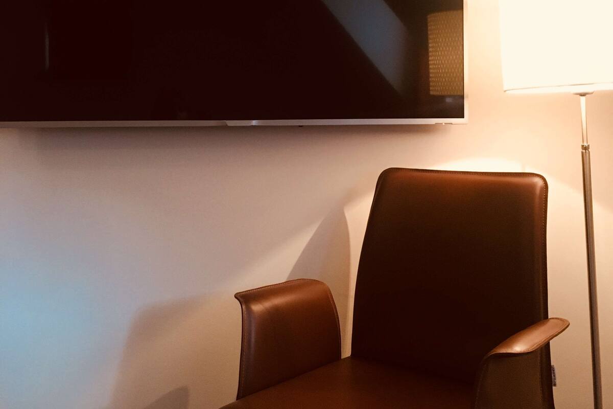Una habitación con televisión de pantalla plana montada en la pared, un sillón de cuero marrón y una lámpara de pie.
